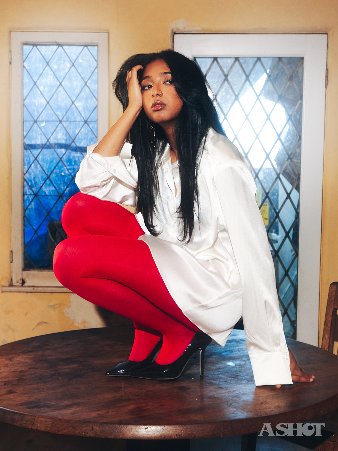 Maitreyi Ramakrishnan in white button up and red tights crouched on a table top and looking longingly away from the camera